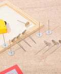 Hella Durable 15 Piece Long Upholstery Nail Set Packed With Heavy Duty Iron Flat Head Push Pins And A Slick Silver Finish displayed on wood.