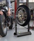 Motorcycle Wheel Balancer And Truing Stand With Adjustable Centering Cones And Truing Pointer For Sport And Dirt Bikes Fits Hollow Axle Wheels Reduces High Speed Vibration For A Smoother Ride; black metal stand balancing motorcycle wheel in workshop.