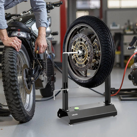 Motorcycle Wheel Balancer And Truing Stand With Adjustable Centering Cones And Truing Pointer For Sport And Dirt Bikes Fits Hollow Axle Wheels Reduces High Speed Vibration For A Smoother Ride; black metal stand balancing motorcycle wheel in workshop.