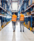 Two workers in orange vests walking through a warehouse aisle with shelves stocked with boxes and goods.
