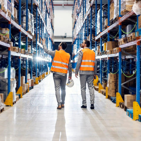 Two workers in orange vests walking through a warehouse aisle with shelves stocked with boxes and goods.