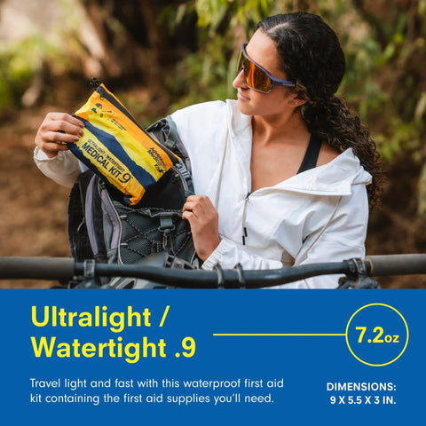 Person holding Ultralight Watertight Medical Kit.9 for up to 4 people, yellow and blue, next to a bike, in outdoor setting.