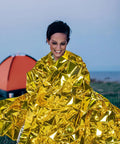 Person wrapped in gold foil from Go-To 6-Piece Survival Emergency Blanket Set, camping scene in background
