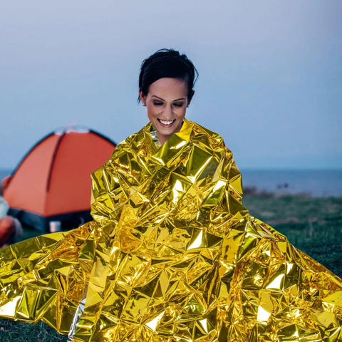 Person wrapped in gold foil from Go-To 6-Piece Survival Emergency Blanket Set, camping scene in background