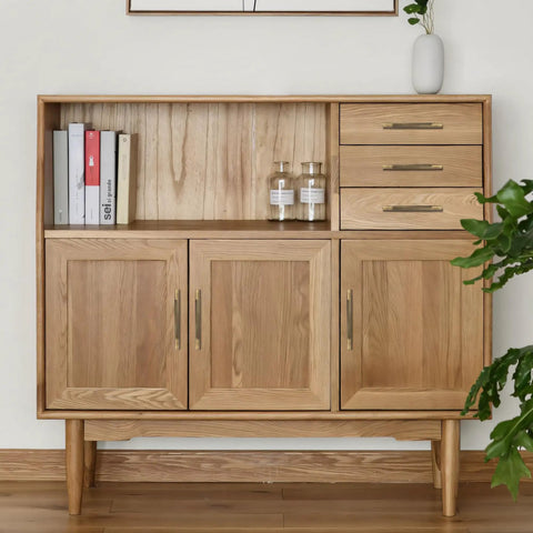 Natural wood cabinet with three doors and two drawers, featuring modern minimalistic design and metal handles, next to a plant.