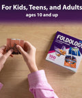 Hands folding paper with Origami Puzzle Game For Brain Teasers Teens And Adults, 100 Puzzles With Five Difficulty Levels visible on table