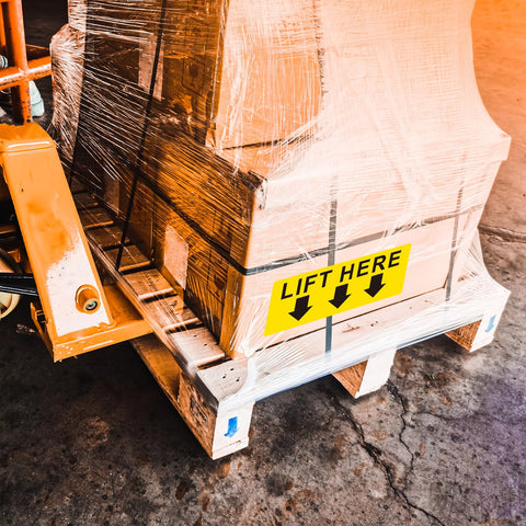 Front view of a pallet with shrink-wrapped boxes and a yellow 5x2 in Lift Here label decal with black arrows