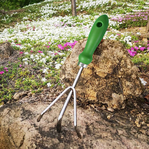 Three Tine Claw Garden Rake with ergonomic handle and stainless steel build resting on rock in the garden