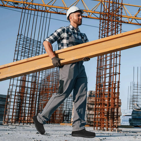 Man wearing Men's Steel Toe Work Shoes Slip Resistant Lightweight Safety Loafers Built For Tough Jobs On Construction And Factory Floors With All Day Comfort And ASTM F2413 18 Impact Protection Black Size 7 carrying wooden beams on construction site.
