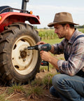 Man using power tool on tractor tire in field, wearing hat, plaid shirt, and denim overalls.