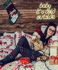 Woman in festive room taking selfie with gifts and 'baby it's cold outside' neon sign on wooden wall.