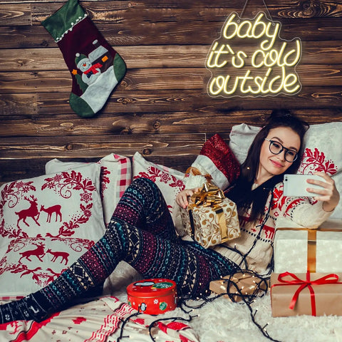Woman in festive room taking selfie with gifts and 'baby it's cold outside' neon sign on wooden wall.
