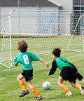 Two kids playing soccer in front of a Backyard Soccer Goal 12 By 6 With Steel Frame Portable Training Net For Kids And Adults Waterproof Ground Stakes Carry Bag Easy Setup For Shooting Practice And Family Fun on grass field.