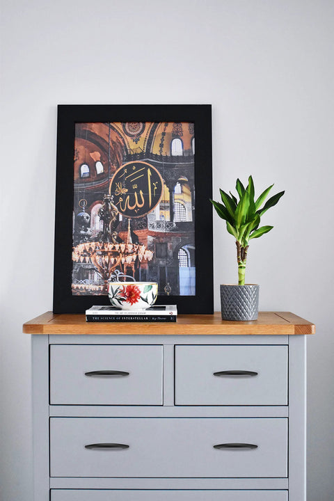Black-framed Islamic art on grey dresser with plant and book