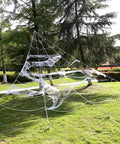 Large outdoor spider web decoration made with ropes and circular anchors on grass, resembling a giant web structure among trees.