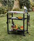 Three Tier Outdoor Grill Cart With Rolling Pizza Oven Table And Stainless Steel Top 26.6 By 16.1 Inches Heavy Duty Steel Stand For Pizza Ovens And Griddles Outdoor Bar Cart With S Hooks For Patio Parties Deck Dining And Camping in a garden setting with various cooking accessories and bottles.