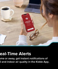 Woman using smartphone to receive alerts from Smart Carbon Monoxide Detector And Indoor Air Quality Monitor with Real Time Alerts No Subscription Needed Plug In With Battery Backup 2 4 GHz Wi Fi Rotating Plug 85 dB Alarm IAQ Insights For Homes.