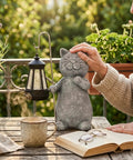 Person touching a Solar Powered Cat Garden Statue With Warm Glow Lantern, Weatherproof Resin Outdoor Decor beside plants on a patio table.