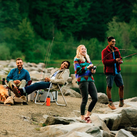 Group of people fishing and sitting by a campfire near a lake, surrounded by rocks and trees