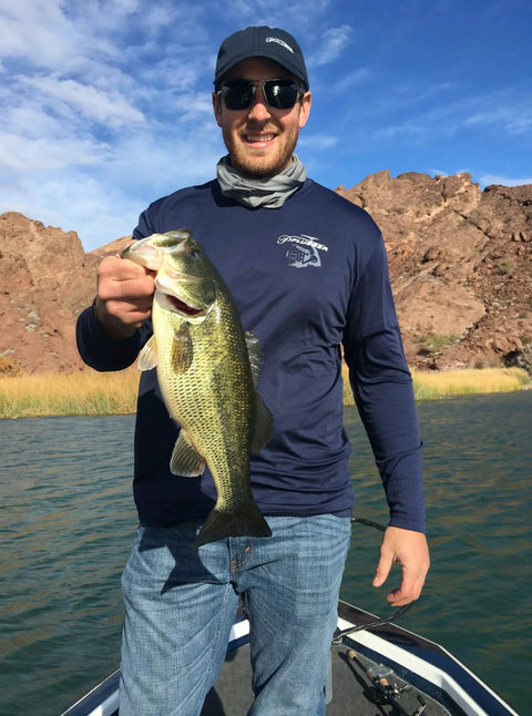 Person standing on a boat holding a largemouth bass in a rocky lake area under blue skies.