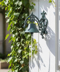 Badass Heavy Duty Cast Iron Outdoor Wall Bell With Slick Vintage Rustic Patina mounted on white wall surrounded by ivy.