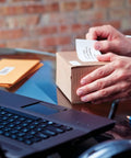 Hands applying a shipping label on a box using the No Sweat Direct Thermal Label Printer With Zero Ink Hassles.