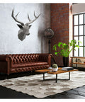 Modern living room with brown leather sofa, geometric deer head wall art, hexagonal rug, and large plant by the window.