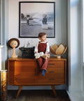 Child sitting on wooden cabinet with doll head and typewriter, black and white photo on wall