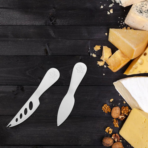 Cheese Knife And Butter Fork Set Stainless Steel With Easy Cutting And Easy Cleanup, next to assorted cheese on dark wood table.