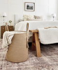 Tall Woven Laundry Basket for Living Room and Bedroom, Boho Farmhouse Round Wicker Blanket Basket for Towels and Clothes, Jute Laundry Hamper with Wooden Bead Tassel, Chic Home Decor 15 by 17.7 Inches beside a neatly made bed in a cozy bedroom setting.