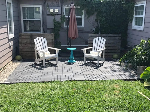 Interlocking Patio Deck Tiles 12x12 Inch Square Composite Plastic Outdoor Flooring For Balcony Porch Backyard All Weather Four Slat Non Slip Tiles Dark Brown Pack Of 27 in a garden setting with two white chairs and a blue table.
