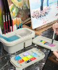 Must-Have Paint Brush Cleaner And Holder With Built-In Palette And Wash Basin For Acrylic, Watercolor, And Water-Based Paints, Compact Portable Organizer In Gray on a workspace with brushes and colorful paints.