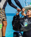 SCUBASak Collapsible Mesh Duffle Bag With Exterior Waterproof Pocket for Dive Gear and Travel on a dock beside the ocean.