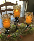 Three frosted glass candle lanterns on a decorative table centerpiece with greenery and flowers, lit with warm glow.
