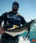 Person holding a large fish on a boat with a fishing rod, wearing Two Conchs Sportfishing shirt under a clear blue sky.