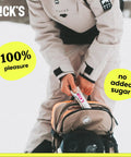 Person placing N!CK'S protein bar into a backpack, emphasizing "100% pleasure" and "no added sugar" as product features.