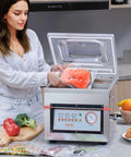 Front view of VEVOR chamber vacuum sealer in stainless steel with clear lid; person loading salmon into a bag.