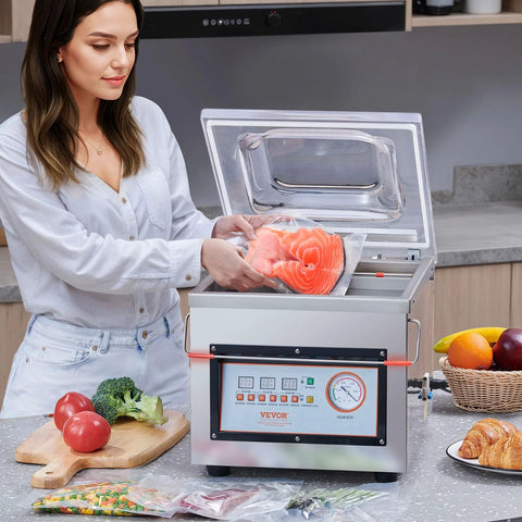 Front view of VEVOR chamber vacuum sealer in stainless steel with clear lid; person loading salmon into a bag.