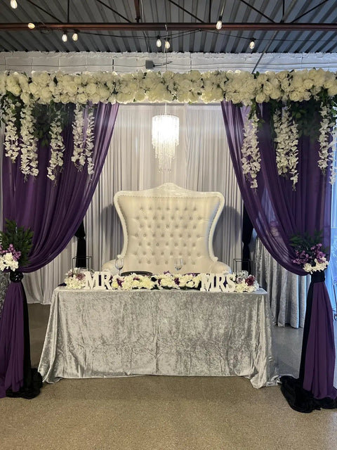 Wedding reception table setup with flowers and purple drapery, featuring a tufted white chair under floral arrangement and string lights.