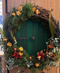 Decorative round green door with floral wreath, colorful flowers, and lush greenery, set against a wooden background.