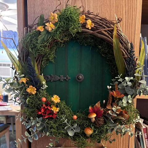 Decorative round green door with floral wreath, colorful flowers, and lush greenery, set against a wooden background.
