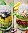 Front view: two clear glass jars with foil-sealed lids and twine, filled with layered salad ingredients.
