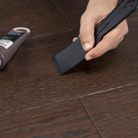 Your Go To Floor Repair Putty In Dark Brown Fast Curing Acrylic Wood Filler For Laminate Vinyl applied with a scraper on a wooden floor.