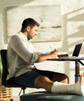 Man using Heavy Duty Adjustable Overbed Table With Ball Bearing Casters to write while seated, with a laptop in view.