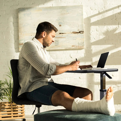Man using Heavy Duty Adjustable Overbed Table With Ball Bearing Casters to write while seated, with a laptop in view.