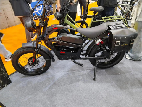 Black electric bike with 20 inch fat tires, displayed at an exhibition, featuring saddle bags and upright handlebars.