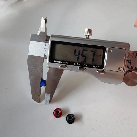 Digital caliper measuring 45.7mm with a blue M3 flange lock nut and red, black nuts nearby. 30 Count Kickass M3 Flange Lock Nuts With Nylon Insert Aluminum Alloy Self Clinching Hex Nut Kit Built For Top Tier FPV Drone And RC Car Rig Upgrades In Sleek Black.