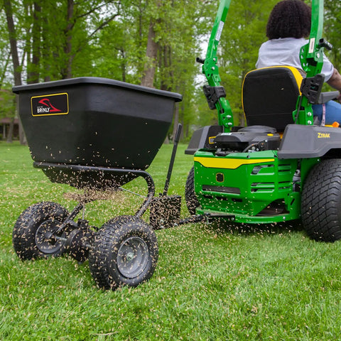 Tow Behind Broadcast Spreader With Universal Hitch 175 Lb Capacity Durable Poly Hopper attached to a mower spreading seeds on grass