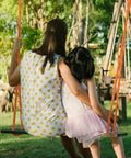 Woman in polka dot dress and child on a wooden swing set outdoors in a park setting with green trees in the background.