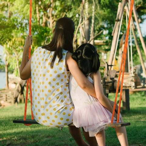 Woman in polka dot dress and child on a wooden swing set outdoors in a park setting with green trees in the background.